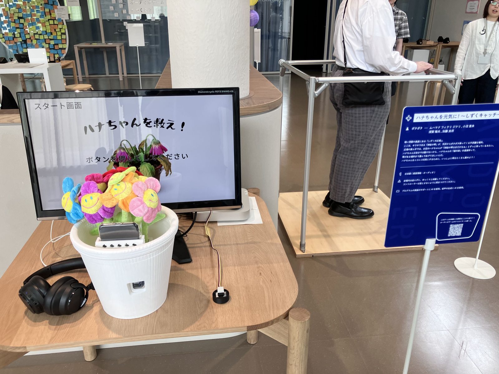 植木鉢のゲーム。「はなちゃんを救え」の文字と枯れた植木鉢がモニターに映し出されている。花のオブジェクトが入った植木鉢とイヤホンが机の上に置かれている。