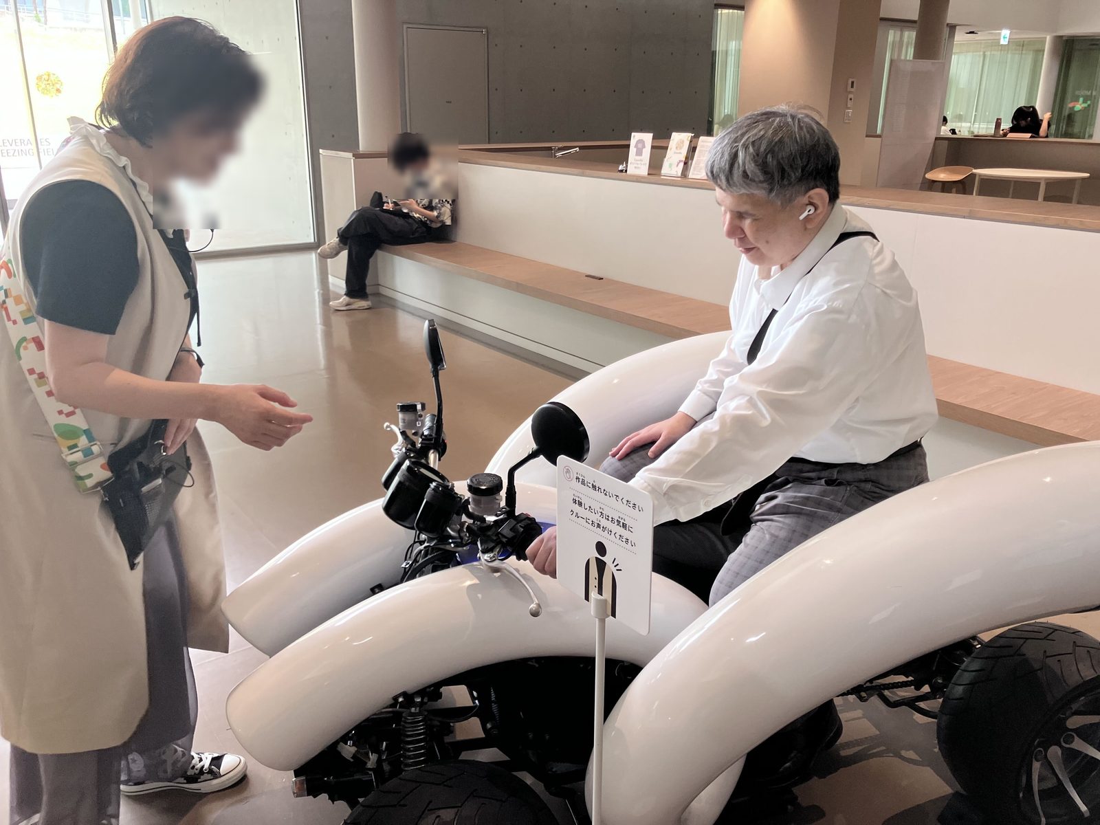 鼓動を伝えるモビリティにまたがった筆者と説明員の様子。乗り物の車体の左右には、白い大きな湾曲した筒が設置されている。